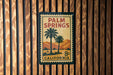 A vintage-style poster of Palm Springs, California, featuring palm trees and mountains, is displayed on a wooden wall.