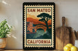 A framed poster of a bridge and mountains, with the text "SAN MATEO CALIFORNIA" on it, is displayed on a shelf next to a wooden cutting board and a potted plant.