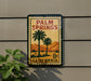 A vintage-style sign for Palm Springs, California, featuring palm trees and mountains, mounted on a wall.