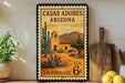 A vintage postage stamp depicting a desert scene with a building and cacti, placed on a shelf next to a wooden cutting board and a potted plant.