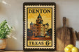 A framed vintage postage stamp depicting a building, likely the Denton Cathedral, in the center of the frame. The stamp is placed on a shelf next to a wooden cutting board and a potted plant.
