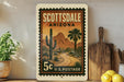 A vintage postage stamp from Scottsdale, Arizona, featuring a desert landscape with a road and cacti, displayed on a shelf next to a wooden cutting board and some lemons.