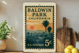 A vintage postage stamp from the United States of California, featuring a sunset landscape and palm trees, is displayed on a shelf next to a wooden cutting board and a potted plant.