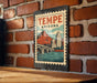 A vintage-style metal sign with the text "TEMPE ARIZONA" and an illustration of a bridge and mountains, mounted on a brick wall.