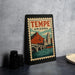 A framed poster of Tempe, Arizona, featuring a mountain and a bridge, is displayed on a countertop next to some black camera lenses.