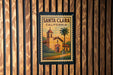 A framed poster of Santa Clara, California, featuring a church and palm trees, is displayed on a wooden wall.