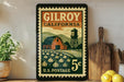 A vintage postage stamp from Gilroy, California, featuring a barn and a water tower, is displayed on a shelf next to a wooden cutting board and a potted plant.
