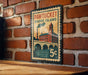 A vintage-style metal sign with a brick wall backdrop, featuring an illustration of a building and a bridge, and the text "Pawtucket Rhode Island" and "5c" indicating the price.