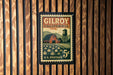 A postage stamp from Gilroy, California, featuring a barn and a field with pumpkins, mounted on a wooden wall.