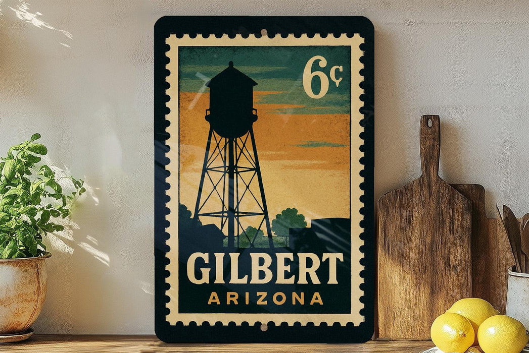 A vintage-style postage stamp depicting a water tower in Gilbert, Arizona, with the text "6¢" and "GILBERT ARIZONA" printed on it. The stamp is displayed on a shelf next to a wooden cutting board and a potted plant.