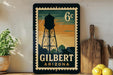 A vintage-style postage stamp depicting a water tower in Gilbert, Arizona, with the text "6¢" and "GILBERT ARIZONA" printed on it. The stamp is displayed on a shelf next to a wooden cutting board and a potted plant.