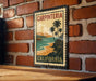 A vintage-style metal stamp depicting a beach scene with palm trees and mountains, mounted on a brick wall.