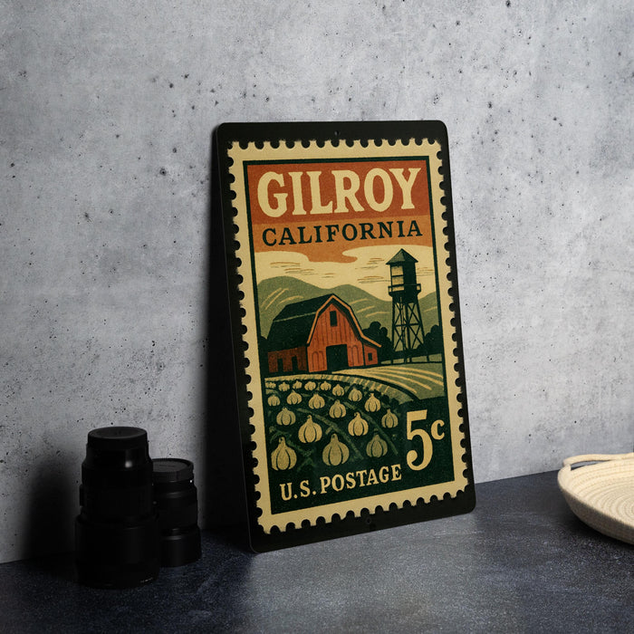 A vintage postage stamp from Gilroy, California, featuring a barn and a water tower, placed on a countertop next to some black camera lenses.