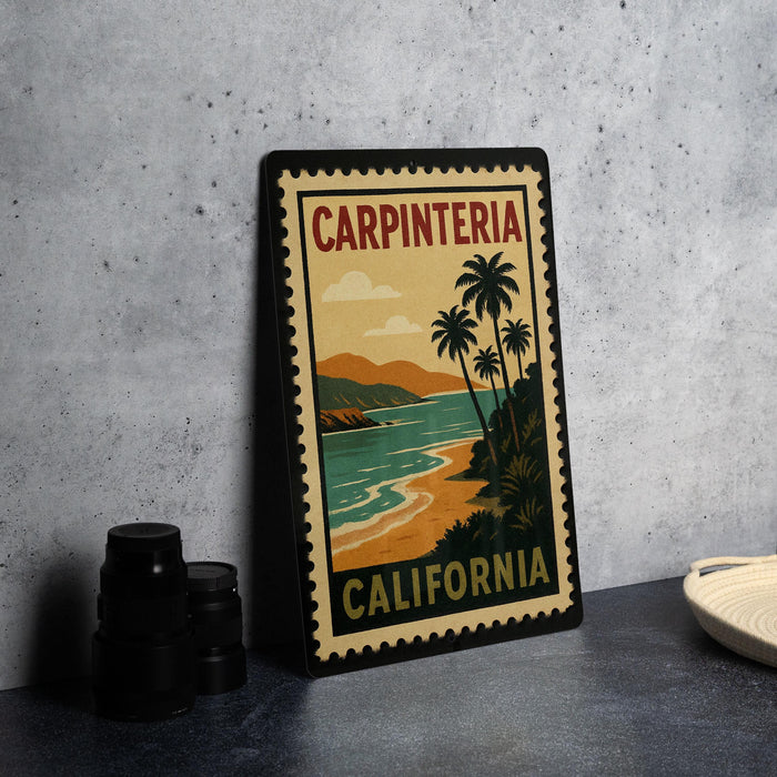 A vintage-style metal stamp with the text "CARPINTERIA CALIFORNIA" and a tropical beach scene, placed on a gray countertop next to some black camera lenses.