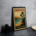 A vintage-style metal stamp with the text "CARPINTERIA CALIFORNIA" and a tropical beach scene, placed on a gray countertop next to some black camera lenses.