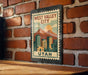 A metal sign with a vintage-style illustration of a city, featuring mountains and buildings, mounted on a brick wall.