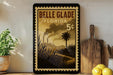 A framed poster of Belle Glade, Florida, with a tropical landscape and industrial smokestacks, is displayed on a shelf next to a wooden cutting board and a potted plant.