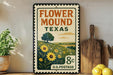 A vintage-style metal sign with a floral design and text, placed on a wooden surface next to a wooden cutting board and some potted plants.