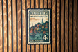 A vintage postage stamp from Massachusetts, featuring a town scene with sailboats and buildings, mounted on a wooden wall.