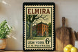 A vintage-style postage stamp from the United States Postal Service, featuring an illustration of a brick building with a tree in the foreground. The stamp is mounted on a wall next to a wooden cutting board and a potted plant.