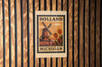 A sign with the text "HOLAND MICHIGAN" and an illustration of a windmill and tulips, mounted on a wooden wall.