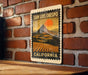 A vintage-style metal sign with a landscape depicting a mountain and a sunset, mounted on a brick wall.