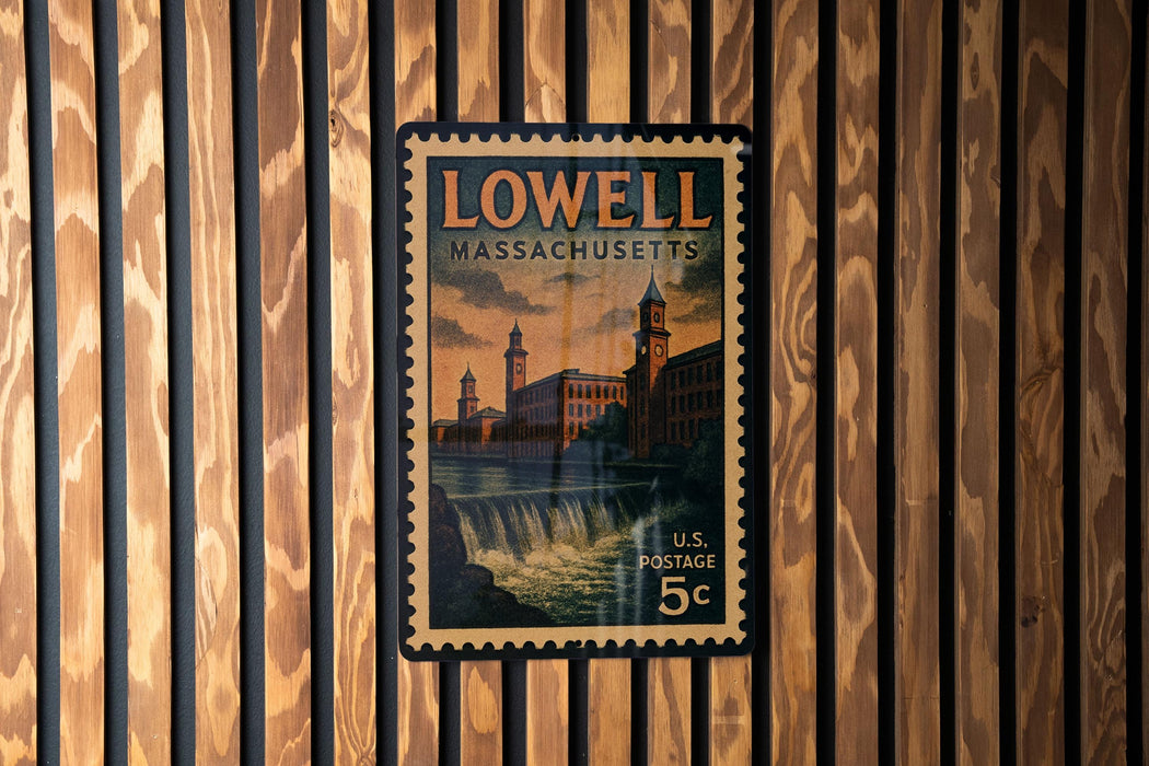 A vintage postage stamp from Lowell, Massachusetts, featuring a building and a waterfall, is displayed on a wooden wall.