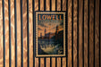 A vintage postage stamp from Lowell, Massachusetts, featuring a building and a waterfall, is displayed on a wooden wall.