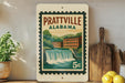 A vintage postage stamp from Alabama featuring a waterfall and buildings, displayed on a shelf next to kitchen utensils and a potted plant.