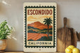 a vintage-style sign for Escondido, California, with a palm tree and a landscape scene, placed on a shelf next to a wooden cutting board and some potted plants.