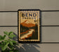 A Bend, Oregon postage stamp is displayed on a wall, featuring a mountain landscape and a river.