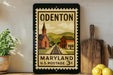 A framed vintage postage stamp from Maryland, featuring a church and train tracks, is displayed on a shelf next to a wooden cutting board and a potted plant.