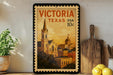 A framed vintage postage stamp from Victoria, Texas, featuring a church and other buildings, is displayed on a shelf next to a wooden cutting board and a potted plant.