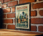 A vintage Boston Massachusetts postage stamp is displayed on a brick wall, featuring a cityscape illustration and the text "U.S. POSTAGE".