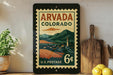 A vintage-style postage stamp from the United States of America, featuring a scenic mountain landscape and a small town, displayed on a shelf next to a wooden cutting board and some lemons.