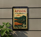 A vintage-style postage stamp from the United States, featuring a mountain landscape and a small town, mounted on a wall.