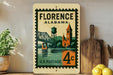 A vintage postage stamp from Florence, Alabama, featuring a bridge and a clock tower, is displayed on a shelf next to a wooden cutting board and some lemons.