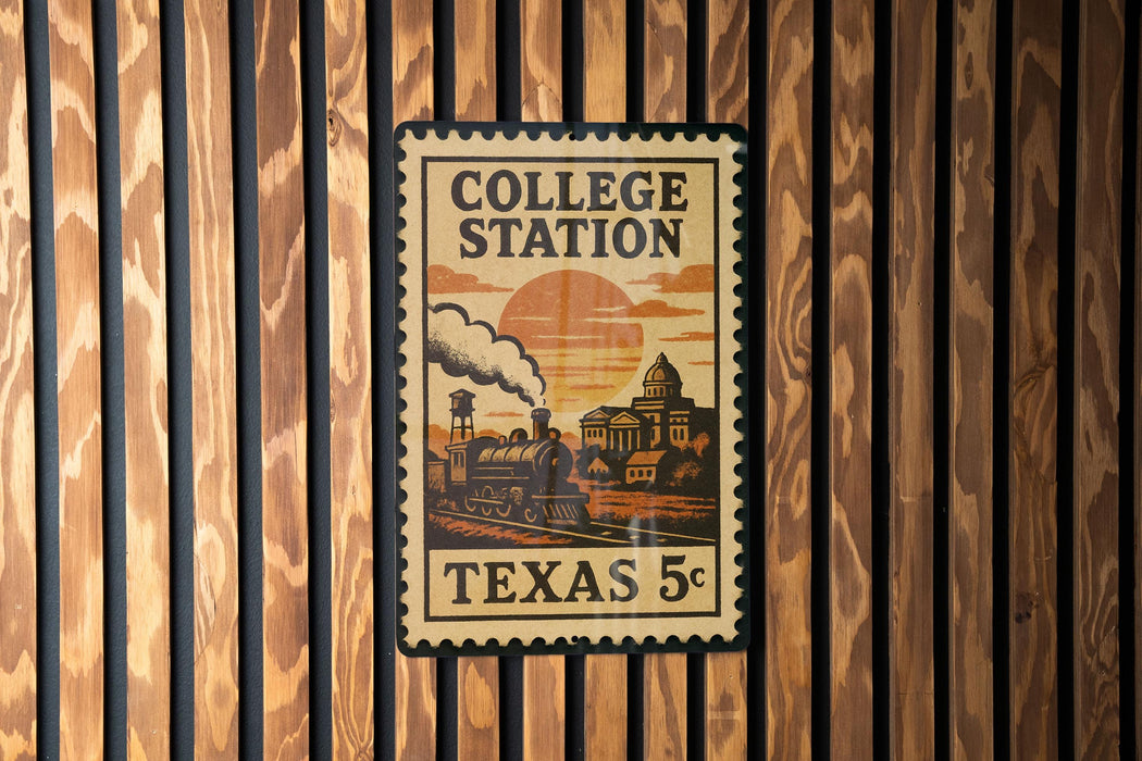 A vintage-style postage stamp depicting a train on a track, with the text "COLLEGE STATION" and "TEXAS 5¢" printed on it, mounted on a wooden wall.