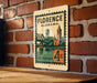 A vintage postage stamp from Florence, Alabama, featuring a bridge and a clock tower, is displayed on a brick wall.