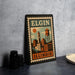 A vintage-style metal sign with the text "ELGIN ILLINOIS" and an illustration of a cityscape, placed on a gray countertop next to some black camera lenses.
