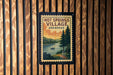 A vintage-style postage stamp depicting a scenic landscape with a river, pine trees, and a small boat, hanging on a wooden wall.