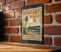 A vintage-style metal sign with the text "NEWPORT BEACH CALIFORNIA" and a palm tree illustration, mounted on a brick wall.