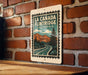 A metal sign with a vintage-style illustration of a road leading to a mountain range, mounted on a brick wall.