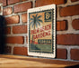 A vintage-style postage stamp featuring a palm tree and the text "Palm Beach Gardens, Florida" is displayed on a brick wall.