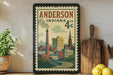 A framed vintage postage stamp depicting a cityscape of Anderson, Indiana, with a tall chimney and buildings, is displayed on a shelf next to a wooden cutting board and a potted plant.