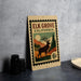 A vintage-style metal sign with a stamp design, featuring a deer and mountains, is displayed on a countertop.
