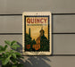 A vintage-style sign for QuINCY, Massachusetts, featuring an illustration of a building and a clock tower.