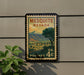 A vintage-style postage stamp from the United States, featuring a scenic landscape with mountains and a town, mounted on a wall.