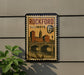 A vintage postage stamp from Illinois, featuring an illustration of a cityscape with a bridge and buildings, mounted on a wall.
