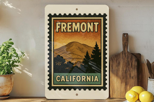 a framed vintage-style stamp with the text "FREMONT CALIFORNIA" and a mountain landscape, placed on a shelf next to a wooden cutting board and some potted plants.